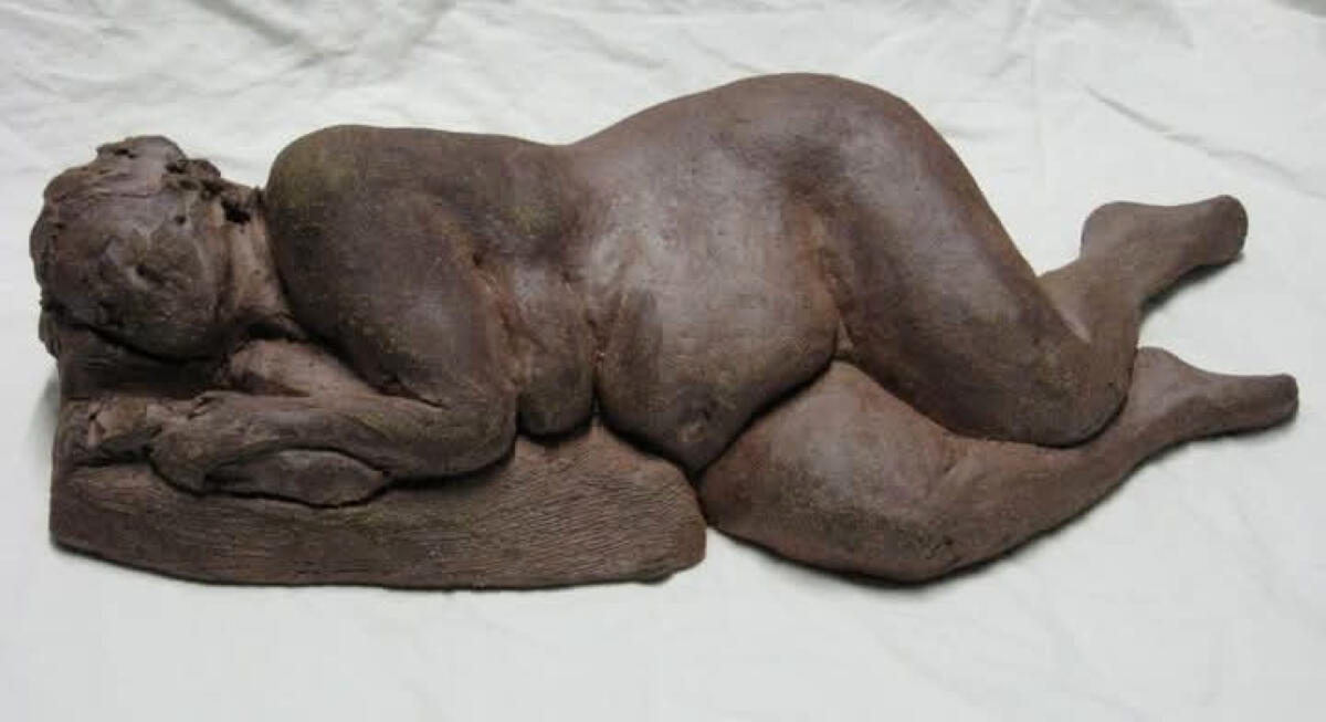 Ceramic sculpture of a reclining nude woman laying on her right side by Shane Brodie. She has a full belly and thighs and wide hips. Facial features are less distinct or detailed. The surface of the piece is a dark matte brown color.