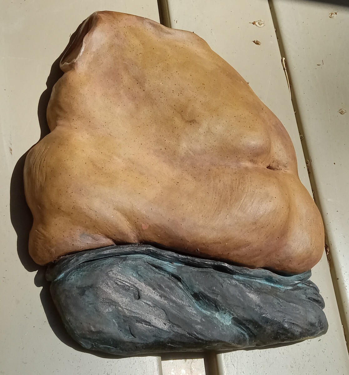 Ceramic bas relief sculpture by Shane Brodie depicting a round belly and right hip with stretch marks. The piece is mostly a speckled golden-brown color. The darker area below the belly has folds highlighted in a light blue.