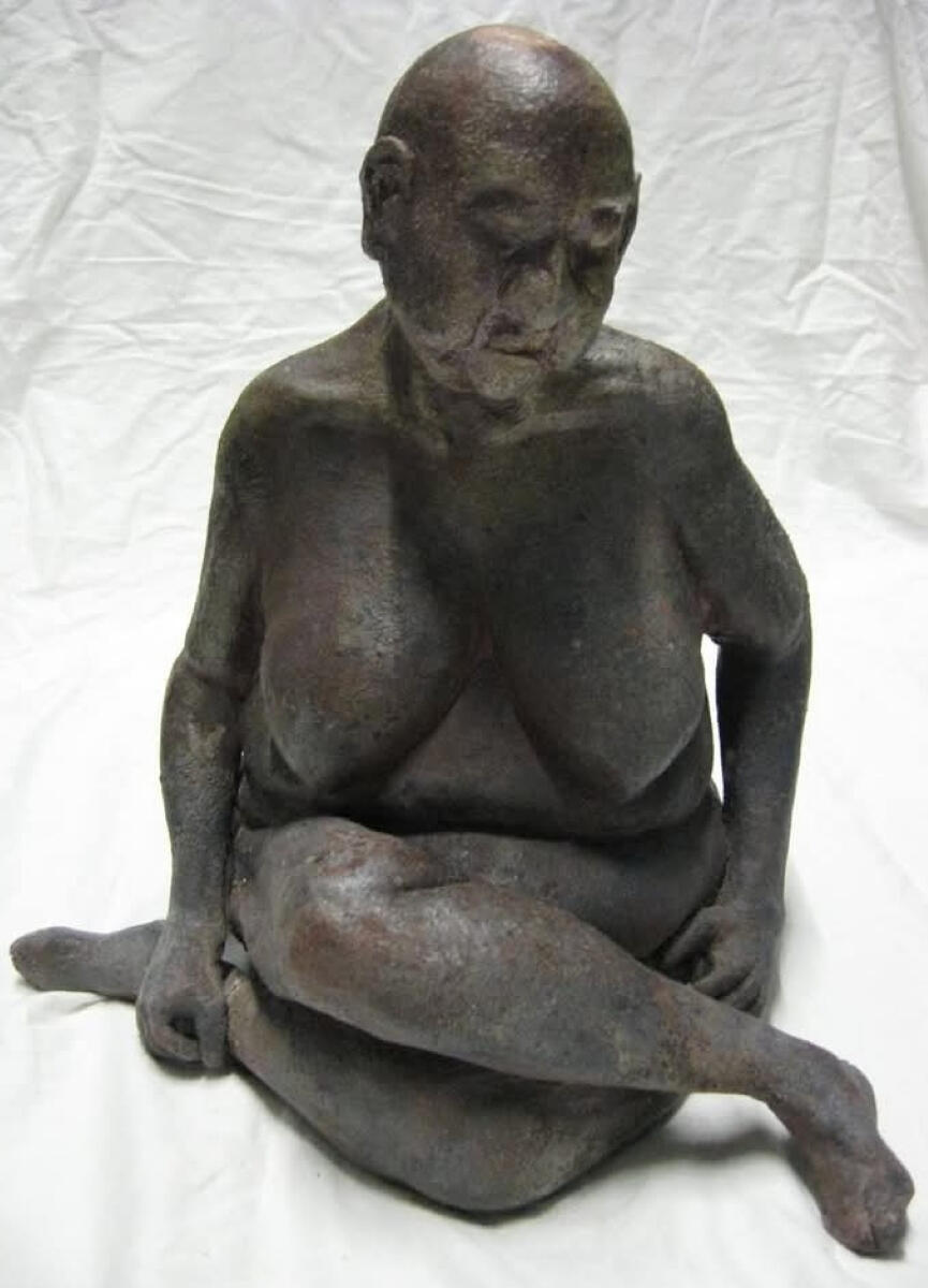 Woodfired ceramic sculpture of a seated older female figure by Shane Brodie. The surface is a mottled dark grey with a blush of red from a heavy reduction environment. The figure is sitting upright with legs crossed and arms down at the sides.