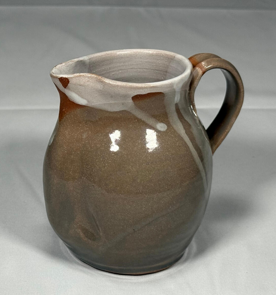 A creamer (a small, short, round pitcher) by Shane Brodie. It is made with stoneware clay with a light green glazed body and a white liner. There are spots of white glaze and unglazed red clay near the rim.