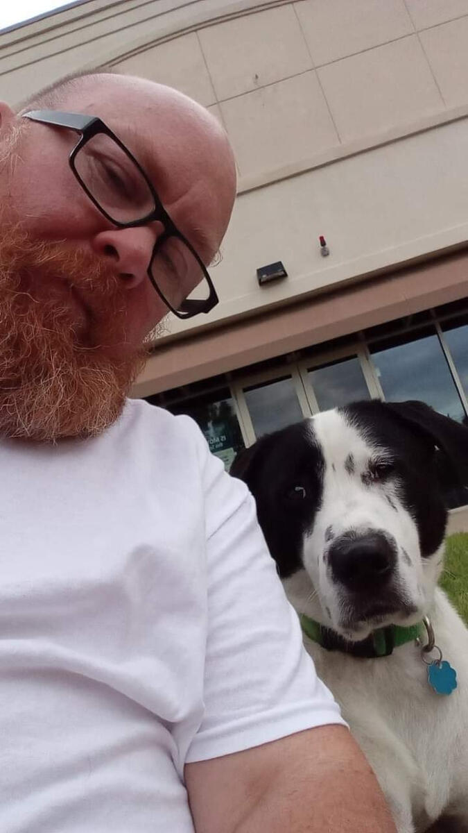 Ceramic artist and activist Shane Brodie with his dog pal, Mr. Cooper. Ceramic artist and activist Shane Brodie on the left. His dog Cooper is on the right. Shane is wearing a white t-shirt and eyeglasses and is a bald, red bearded trans man. Cooper is a 70-pound, mixed breed dog with mostly white fur and black spots.